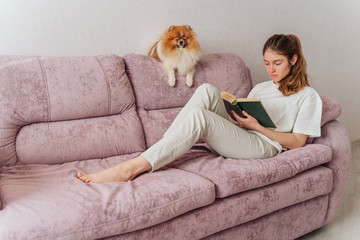 the girl is resting on the sofa reading a book. a furry companion dog is resting nearby. a woman reads an interesting book