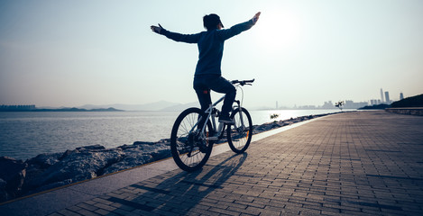 Female cyclist hands free cycling riding bike with arms outstretched in the coasts sunrise - obrazy, fototapety, plakaty