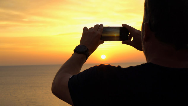 Travel concept male hand holding a smartphone for taking a photo of the sunset over the sea