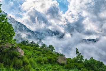 Majestic view of high mountains hidden by clouds and fog from a green cliff.