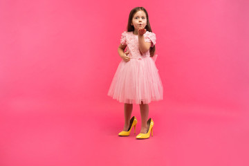 Fashionable cute young girl with long brunette hair, in stylish dress wearing big mother`s shoes, sending kiss to camera isolated on pink background. Having fun of joyful kid. Place for text.