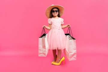 Happy lovely moments of shopping time with cute little girl in dress standing in mother`s big shoes with white packages in hands isolated on pink background