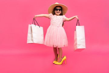 Happy lovely moments of shopping time with cute little girl in dress standing in mother`s big shoes with white packages in hands isolated on pink background