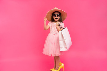 Obraz na płótnie Canvas Happy lovely moments of shopping time with cute little girl in dress standing in mother`s big shoes with white packages in hands isolated on pink background