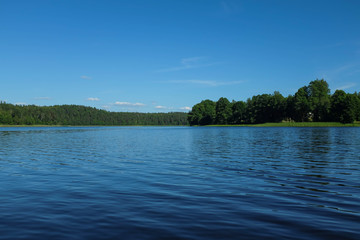 Scenic landscape of forest lake. Lake shore with green trees. Sunny summer day. Blue sky.