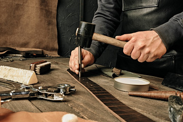 Working process of the leather belt in the leather workshop. Man holding hammer. Tanner in old tannery. Wooden table background. Close up man arm. Maintenance concept. Goods production. - Powered by Adobe