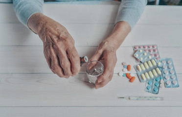 Senior hands with medicine bottle putting drops in a glass, pills, drugs, thermometer on a table. Woman wrinkled hands, colorful tablets, wooden background. Health care for the elderly, old age.