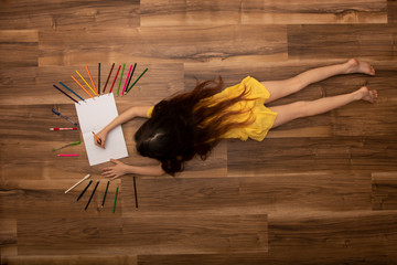 Top view of charming little girl drawing using colorful pencils while lying on the floor in her room at home