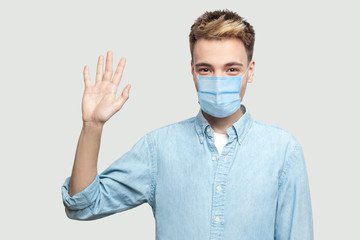 Portrait of happy excited handsome young man with surgical medical mask in blue shirt standing, looking at camera and waving his hand to greeting. indoor studio shot on grey background copy space.
