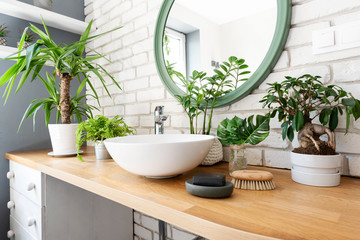 Interior of bathroom with a lot of plants on wooden counter and round mirror on white brick wall....