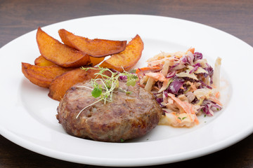 A burger with baked potatoes, cabbage and a salad in a ceramic plate on a wooden table