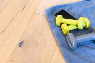 Fitness at home, equipment and tools for gym at home. Green dumbbell with towel, mobile phone on wooden parquet floor. Background with copyspace for sport activity at home with accessories.