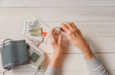 Senior hands with medicine bottle putting drops in a glass, pills, tonometer, thermometer on a table. Woman wrinkled hands, colorful tablets, blood pressure cuff. Elderly health care, old age.