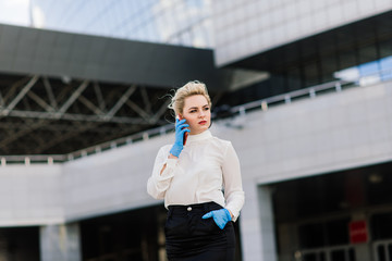 Portrait of a young businesswoman with phone, notebook, tablet, coffee outdoors. Blonde girl in rubber blue gloves.