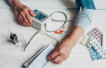 Senior wrinkled hands with blood pressure measure cuff, medicine bottle, glass, colorful pills, thermometer on a table. Old woman checking blood pressure on a tonometer. Elderly health care, old age.