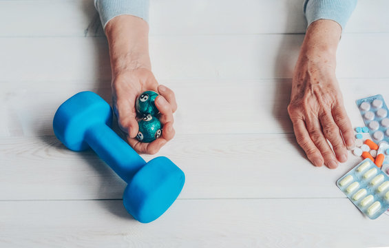 Senior wrinkled hand pushing back pills and thermometer, dumb-bell on a table. Another old woman hand with chinese health exercise baoding balls. Elderly health care, old age, healthy living concept.