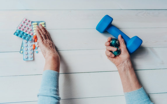 Senior wrinkled hand pushing back pills and thermometer, dumb-bell on a table. Another old woman hand with chinese health exercise baoding balls. Elderly health care, old age, healthy living concept.