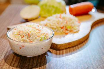 Coleslaw salad in glass bowl on wooden table and chopped cabbage, onion and carrot on cutting board on background