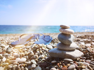 Pyramid of sea stones on pebbles of the sea shore. Two blue slippers  on the beach on a sunny warm summer day on the the blue sea background. The concept of relaxation and long-awaited vacation. 