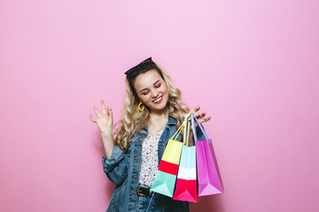 Portrait of a young blond girl holding shopping bags and showing gestures on pink background