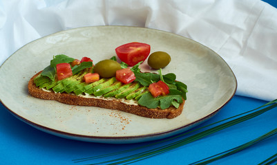 Avocado toast of rye bread with sauce, tomatoes, arugula on large grey plate on blue background. Vegetarian useful breakfast.