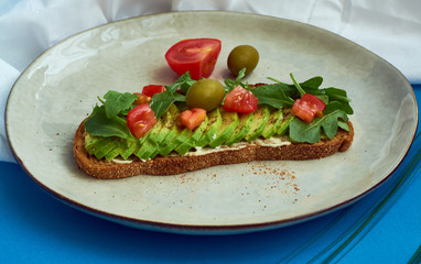 Avocado toast of rye bread with sauce, tomatoes, arugula on large grey plate on blue background. Vegetarian useful breakfast.