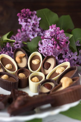 Obraz na płótnie Canvas Bouquet of lilac flowers and chocolate candies with filling and nuts, chocolates with nuts on a plate, background. Sweets made of black, milk and white chocolate with hazelnuts, almonds and cashews
