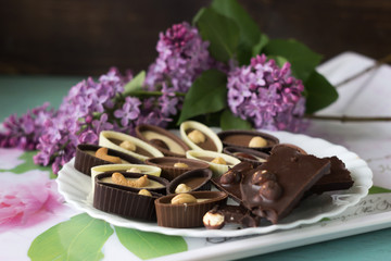 Bouquet of lilac flowers and chocolate candies with filling and nuts, chocolates with nuts on a plate, background. Sweets made of black, milk and white chocolate with hazelnuts, almonds and cashews