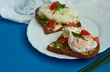 Poached eggs on sandwich of rye bread with sauce, tomatoes and arugula in white plate on blue background.