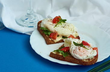 Poached eggs on sandwich of rye bread with sauce, tomatoes and arugula in white plate on blue background.