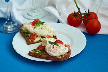 Poached eggs on sandwich of rye bread with sauce, tomatoes and arugula in white plate on blue background.