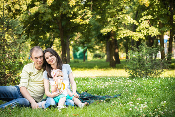 Fototapeta premium Happy young parents sitting in park with their small baby daughter on knees on summer clear day. Happy family time together concept