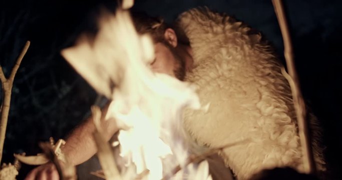 Man dressed in a sheepskin warming himself at a campfire in a close up on the flames as he places his hands close to the warmth.