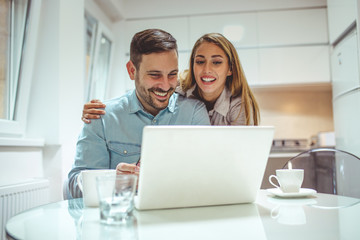 Shot of a young woman hugging her husband while he uses a laptop at kitchen. Portrait of cheerful couple using laptop together while sitting at home. Couple having fun at home
