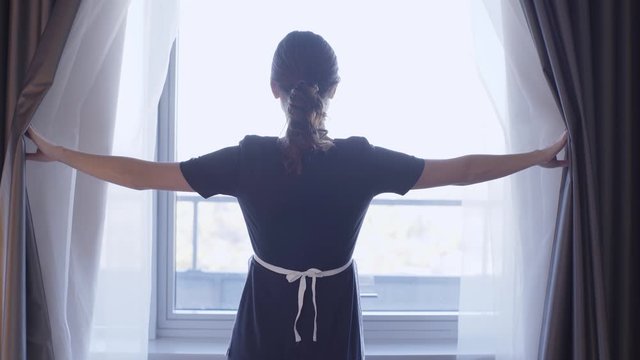 back view of young maid opening dark curtains in hotel room