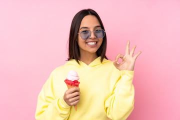 Young brunette girl holding a cornet ice cream over isolated pink background showing an ok sign with fingers