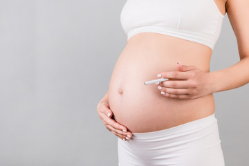 Close up of pregnant woman in white underwear with a cigarette against her belly at gray background. Dangerous risk for unborn baby