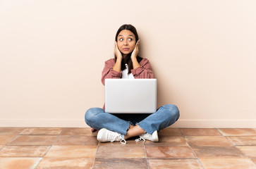 Young mixed race woman with a laptop sitting on the floor frustrated and covering ears