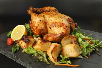 Whole roasted grilled chicken poultry bird with baked potato vegetable salad tomato lemon on black slate stone cutting board black background