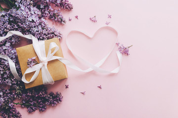 Lilac flowers, gift box and heart ribbon on pink paper, stylish flat lay with copy space. Floral greeting card. Happy mothers day concept. Purple lilac flowers bouquet with craft present box