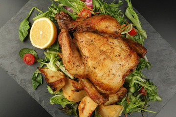 Whole roasted grilled chicken poultry bird with baked potato vegetable salad tomato lemon on black slate stone cutting board black background