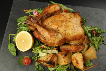 Whole roasted grilled chicken poultry bird with baked potato vegetable salad tomato lemon on black slate stone cutting board black background