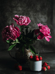Obraz na płótnie Canvas A bouquet of peonies in a vintage vase. The flowers are pink in colour. Next to it is a mug of strawberries. Summer still life on a dark background.