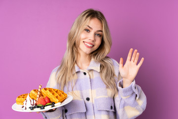 Teenager Russian girl holding waffles isolated on purple background saluting with hand with happy expression