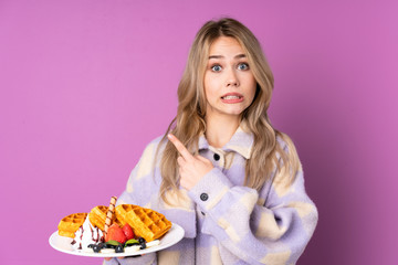 Teenager Russian girl holding waffles isolated on purple background frightened and pointing to the side