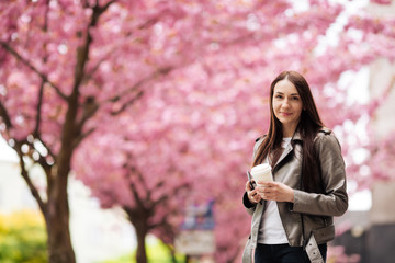 Girl in stylish outfit smiling and holding coffee on sakura background. Portrait of woman in jacket with cup of tea