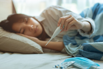 Asian teen infected with Covid-19 flu sick lying in bed due to a Corona virus pandemic, anxiously measuring check body temperature with digital thermometer. - obrazy, fototapety, plakaty