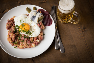 Norddeutscher Hamburger Labskaus mit Rindfleisch Corned Beef, Rote Beete, Kartoffeln, Saure Gurken, Spiegelei, Rollmops und Bier auf Holztisch