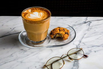 Coffee latte in a glass cup, a cookie and old eye glasses on a marble table