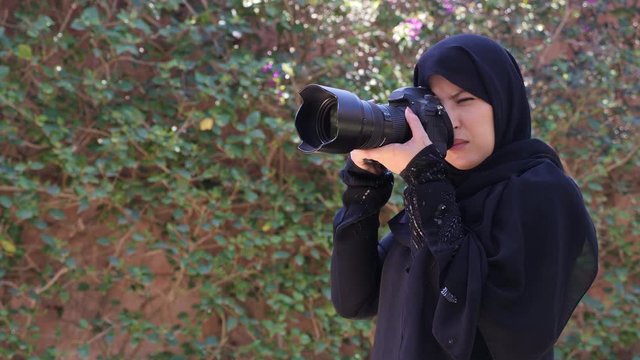 A beautiful Moroccan Arab Muslim woman with a DSLR camera filming photographing in a garden - turning the camera away from the viewer and up. Islamic emancipation. Real-time footage.